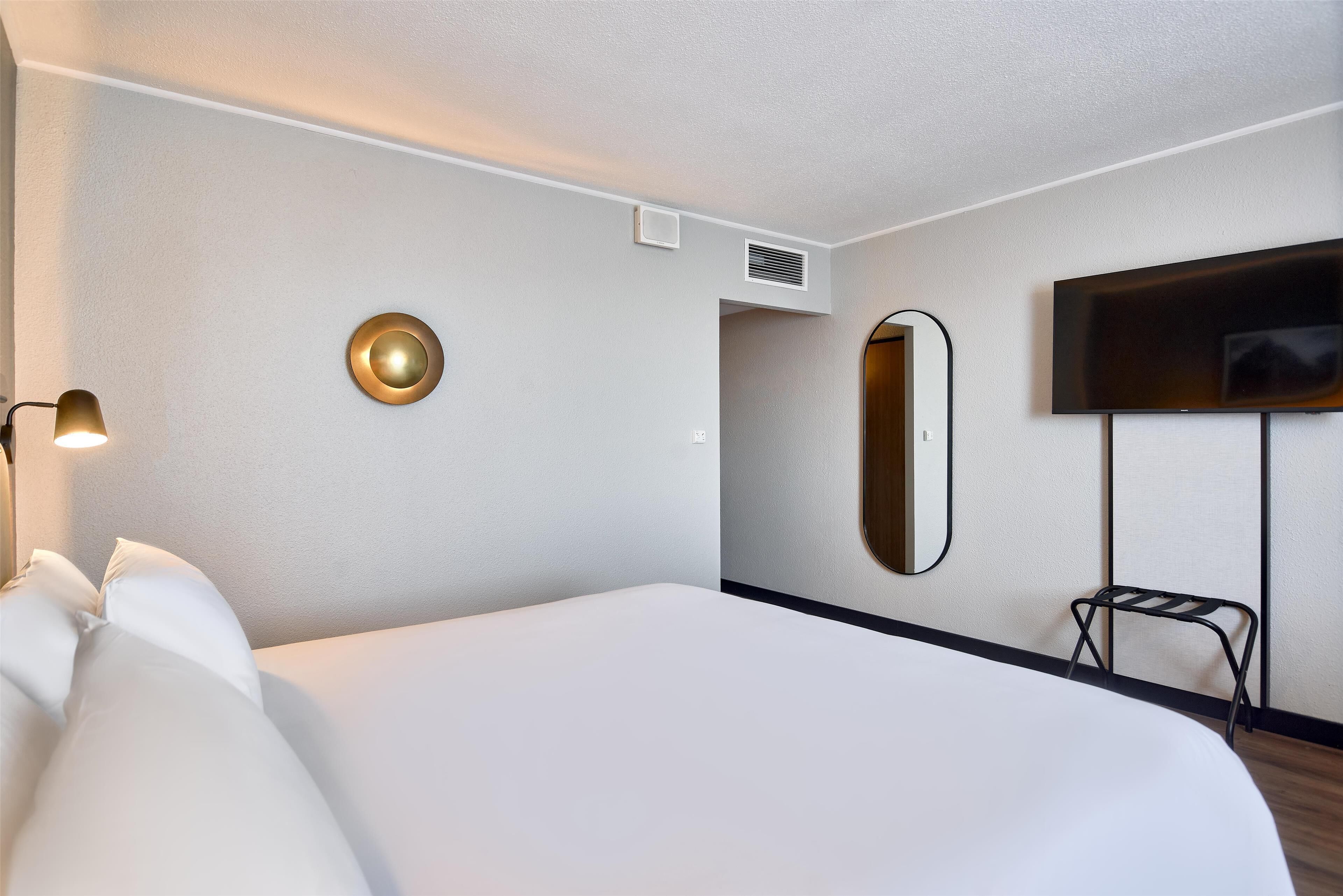 A hotel room with a large bed, a wall-mounted TV, a round mirror, a small stool, and a wall-mounted lamp.