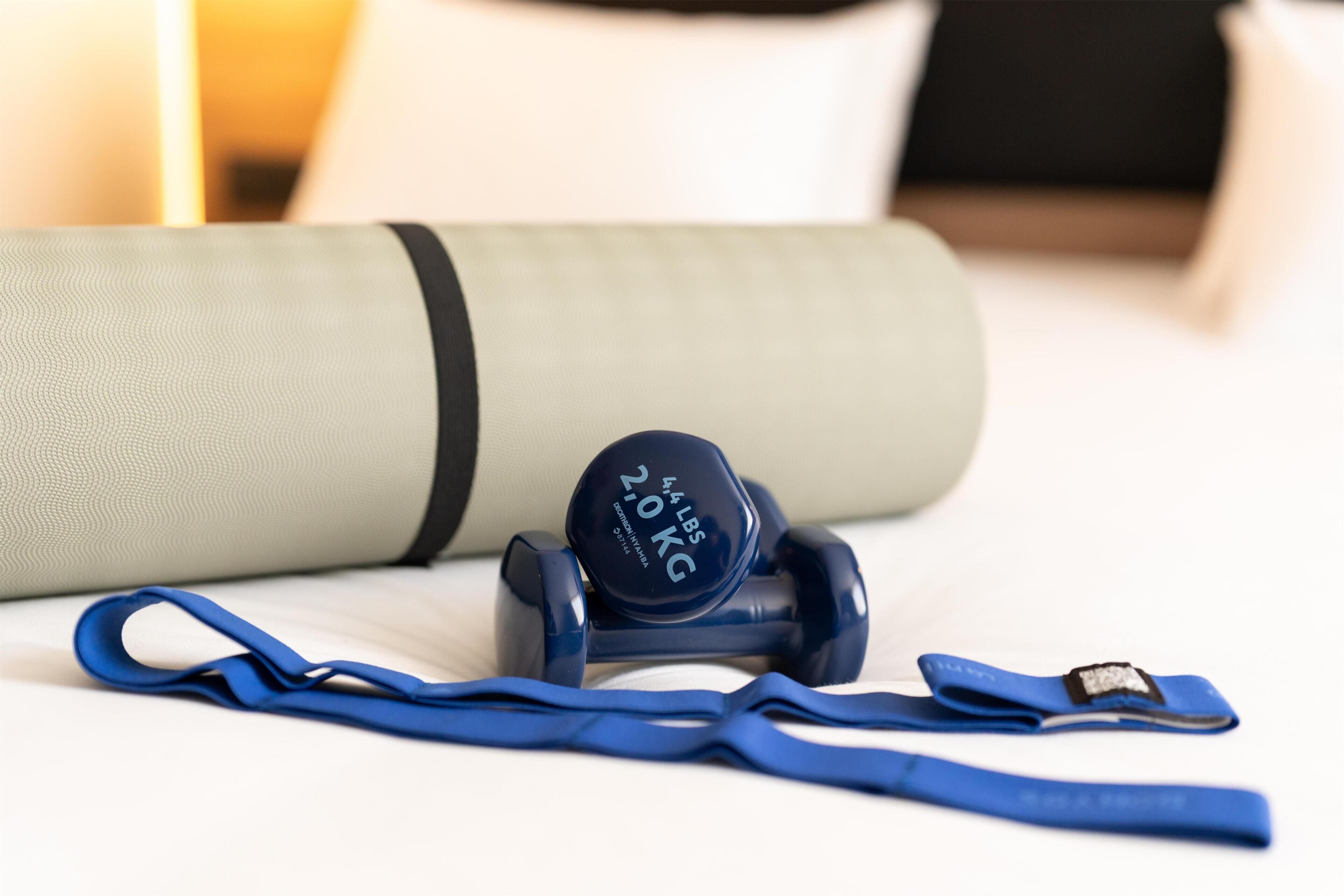 A bed with a rolled-up yoga mat, a blue resistance band, and a weight plate labeled '4.5 LBS 2.0 KG'.