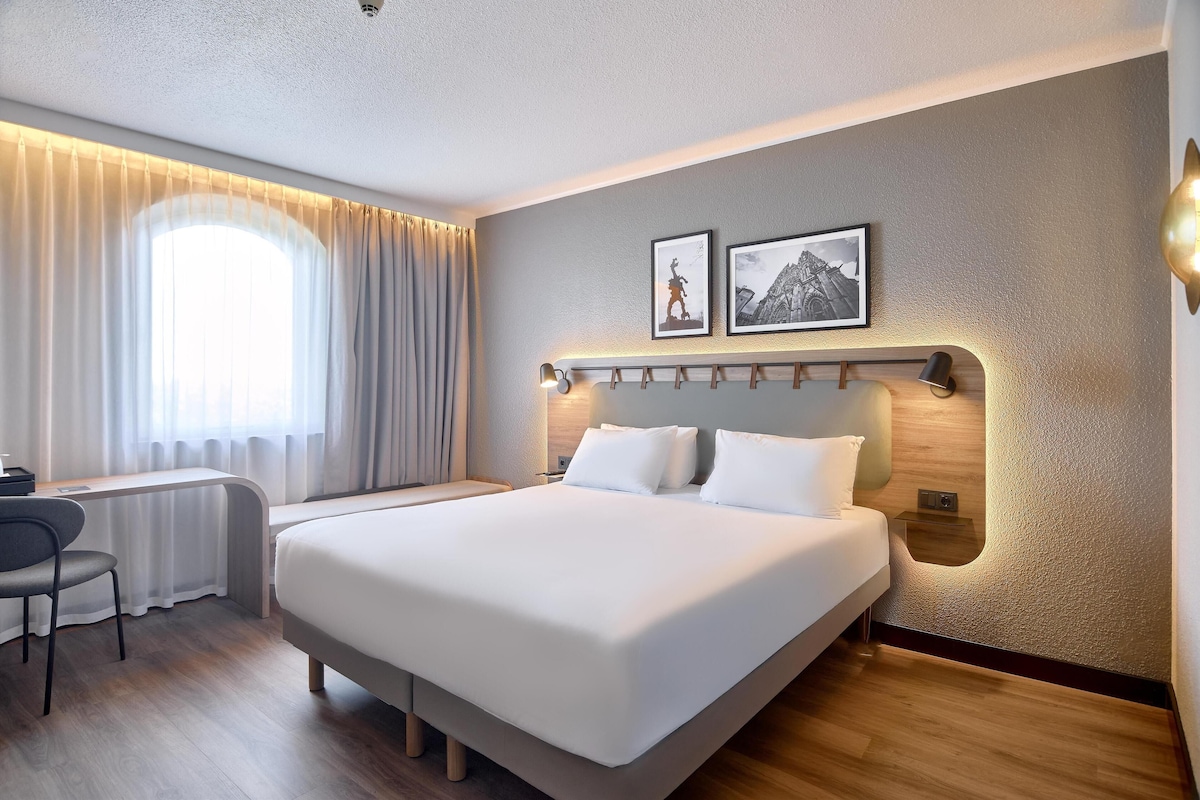 Une chambre d’hôtel moderne avec un grand lit, un bureau et une chaise. Deux tableaux encadrés ornent le mur, et une fenêtre est agrémentée de rideaux.