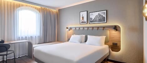 Una habitación de hotel moderna con una cama grande, un escritorio y una silla. Hay dos cuadros en la pared y una ventana con cortinas.