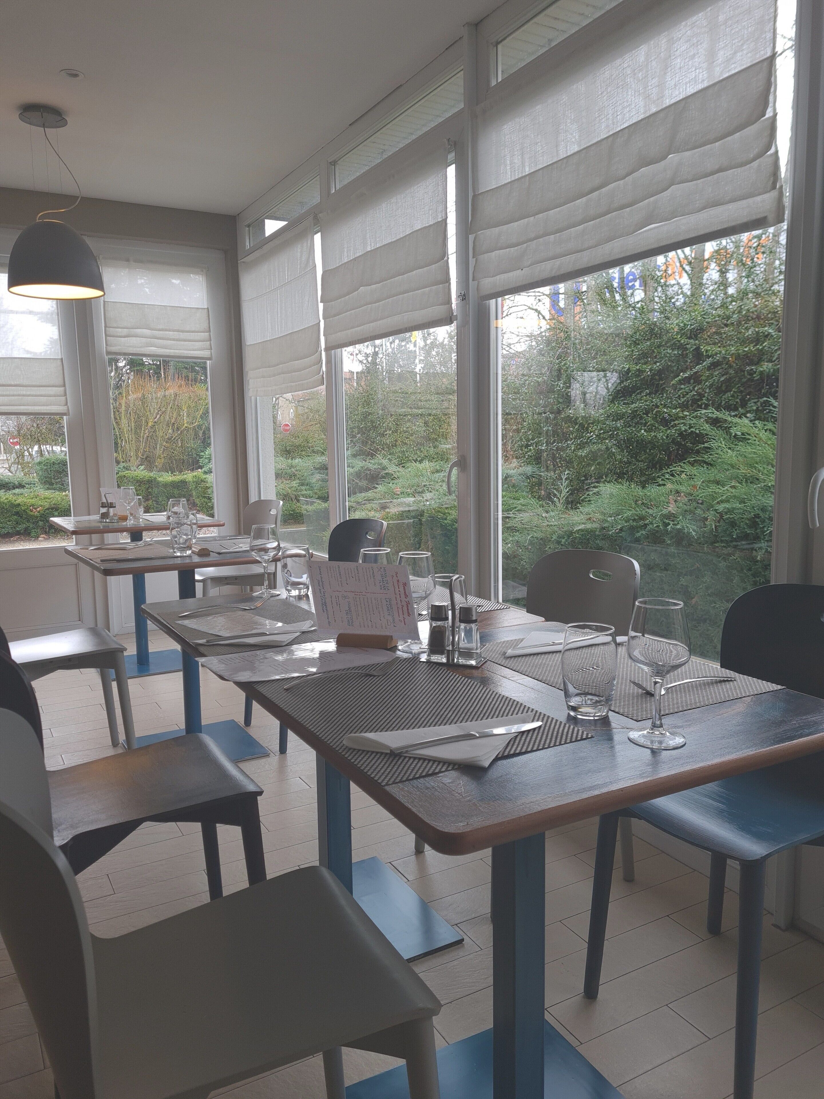 Un restaurante con mesas dispuestas para comer, ventanas amplias con persianas y vistas a espacios verdes exteriores.