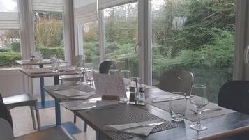 Un restaurante con mesas dispuestas para comer, ventanas amplias con persianas y vistas a espacios verdes exteriores.