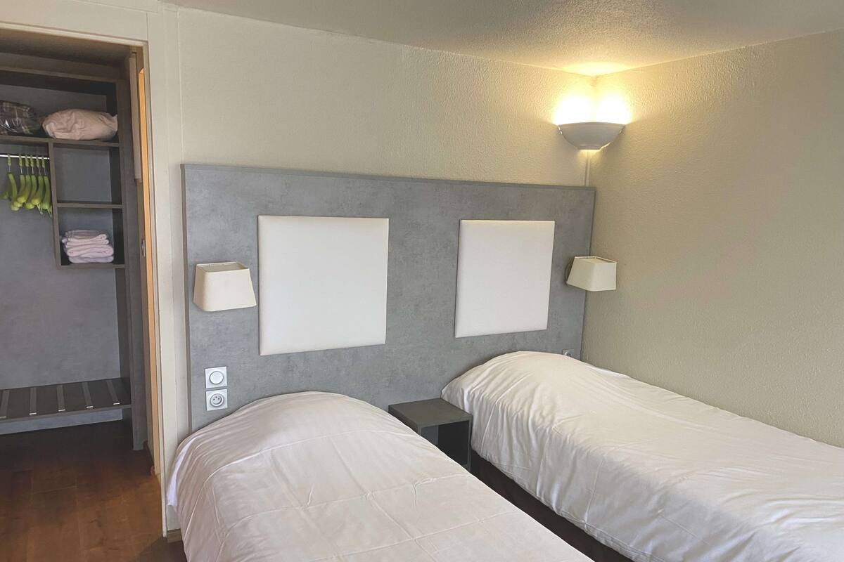 A hotel room with two single beds, a wardrobe with hanging clothes, and a wall-mounted lamp.