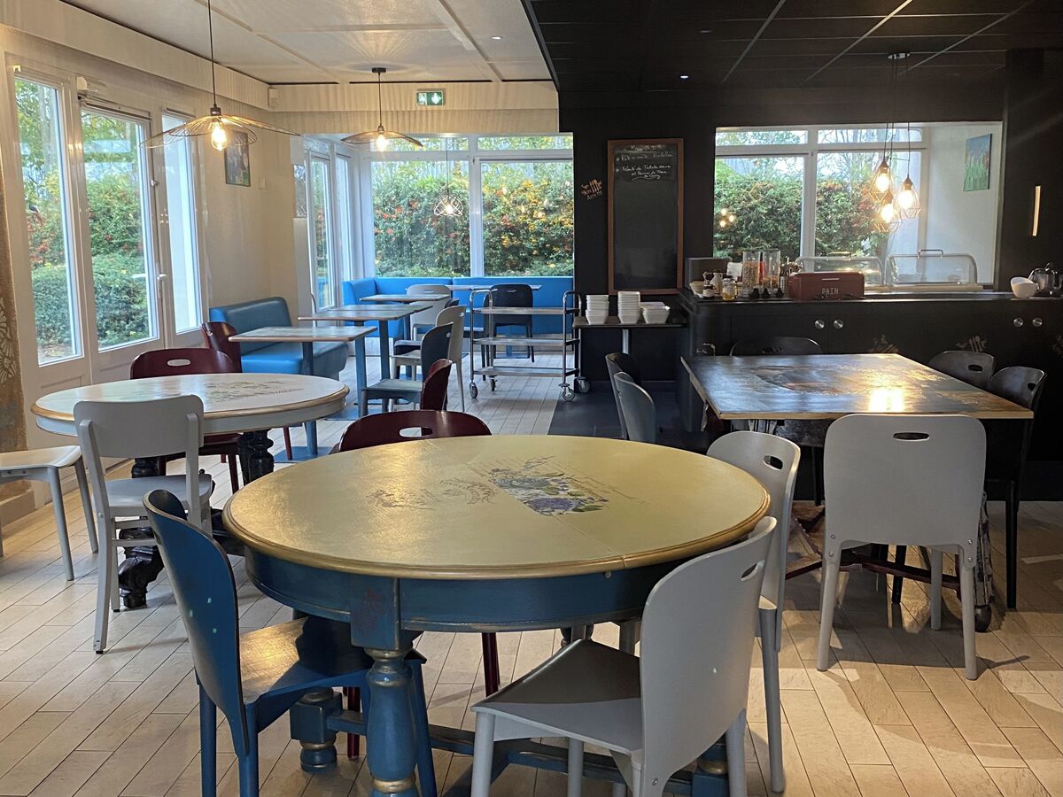 A restaurant with round tables and chairs, large windows, and a view of greenery outside.