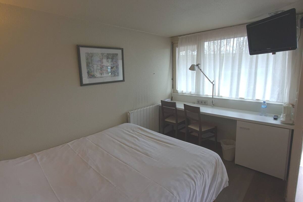 A room with a bed, a desk, a chair, a window with curtains, a picture on the wall, and a television mounted on the wall.