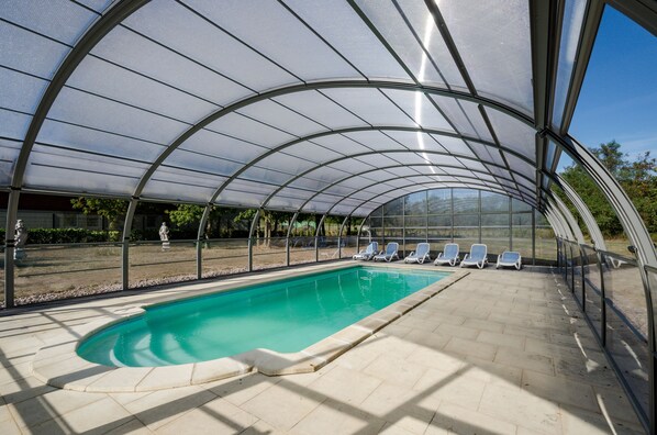 A covered swimming pool with clear glass walls and a tiled floor, surrounded by lounge chairs.