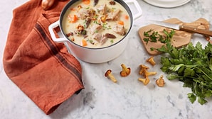 Un tazón de sopa cremosa con verduras y carne, un molinillo de pimienta de madera, una tapa blanca, una tabla de cortar de madera con un cuchillo, y perejil fresco.