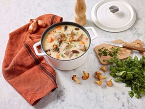 A pot of creamy soup with vegetables and meat, a wooden pepper grinder, a white lid, a wooden cutting board with a knife, and fresh parsley.