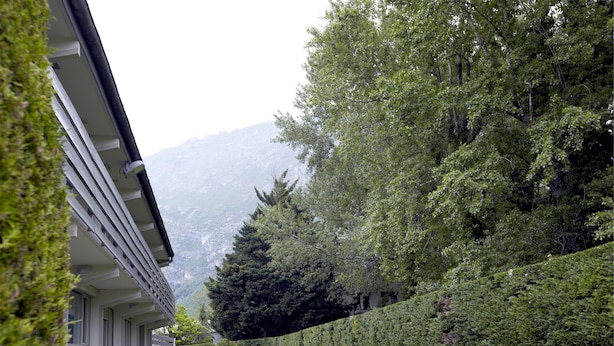 Un edificio con un espacio al aire libre cubierto, rodeado de vegetación cuidada y montañas al fondo.