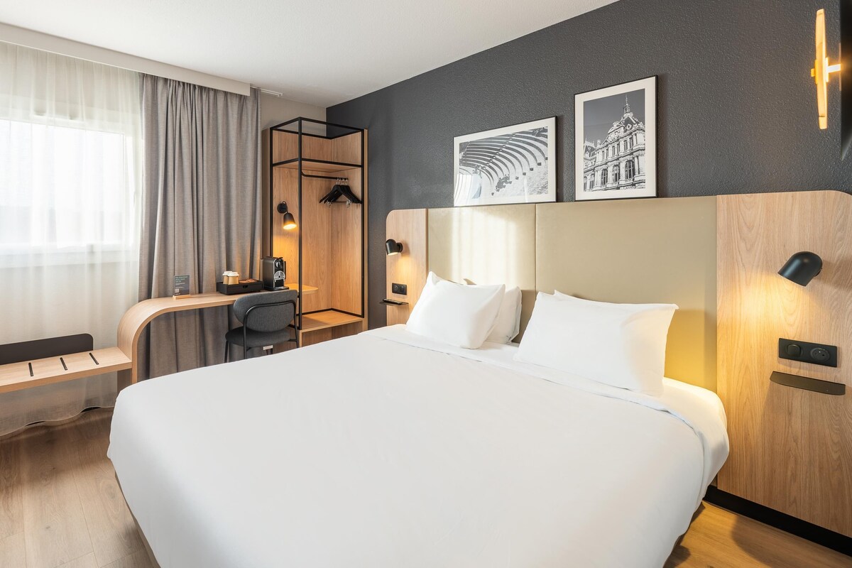A hotel room with a large bed, a desk with a chair, a small seating area, and artwork on the wall.