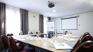 Un salón de conferencias con una larga mesa, sillas rojas, tarjetas de identificación, botellas de agua y una pantalla de proyector.