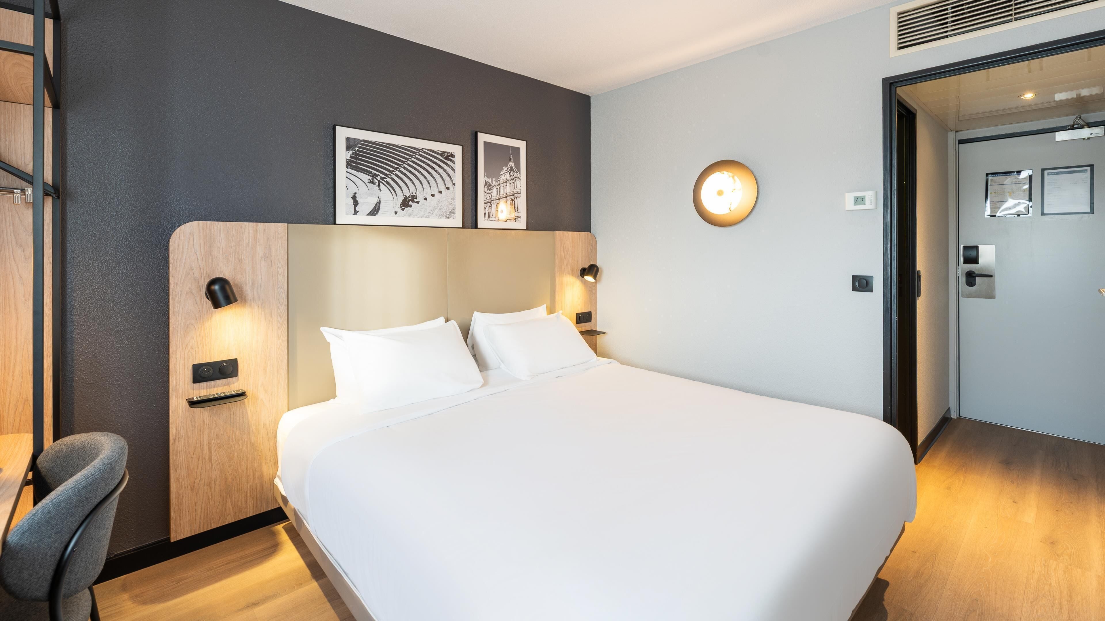 A hotel room with a large bed, a desk, and a chair. There are two wall-mounted lamps and a clock on the wall.