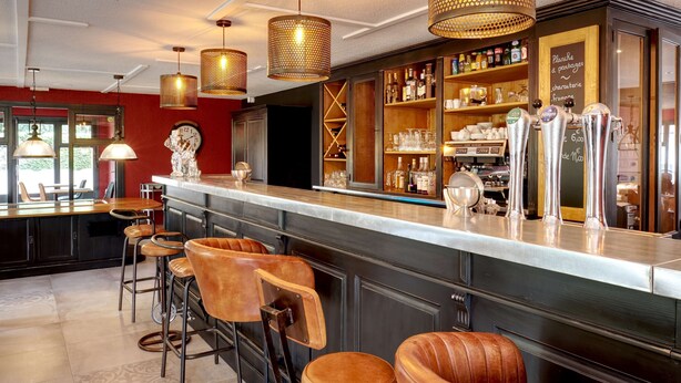 A bar area with stools, a counter, and a shelf with bottles. - Kyriad Lille Est - Hem (Hem)