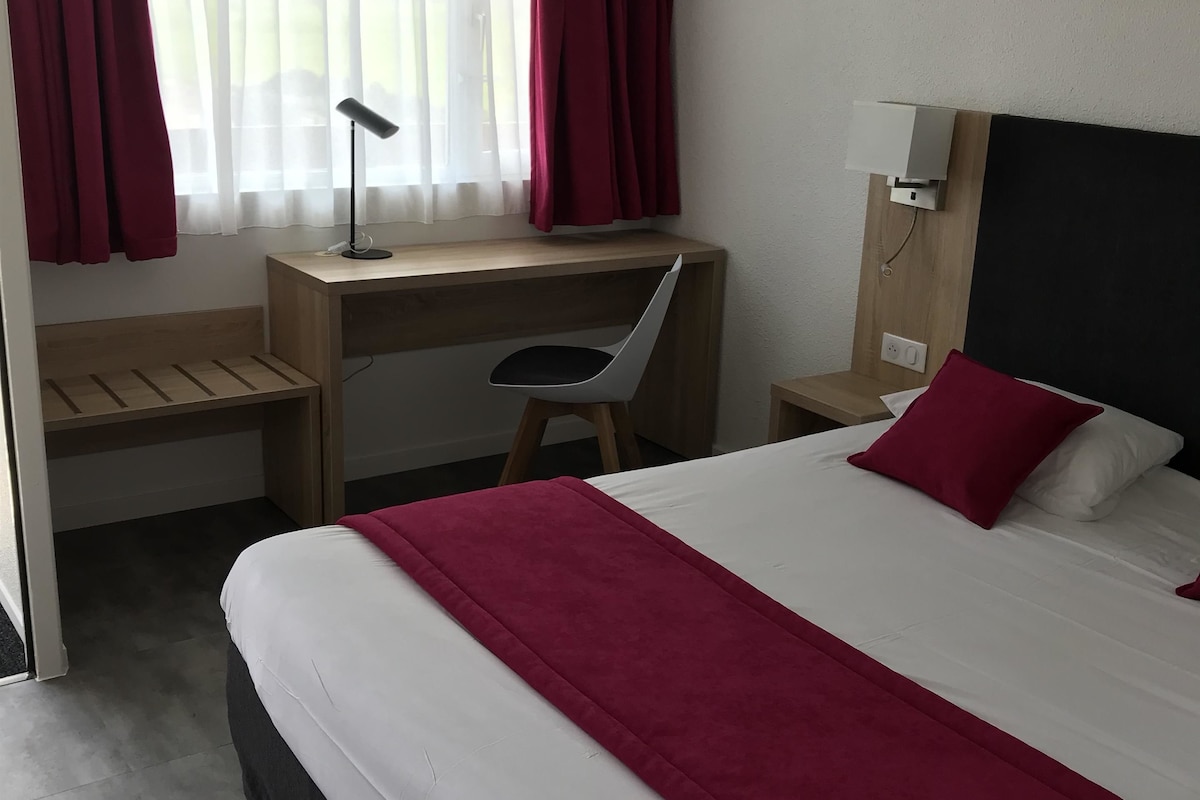 A hotel room with a bed, a desk with a chair, a lamp, and a window with curtains.