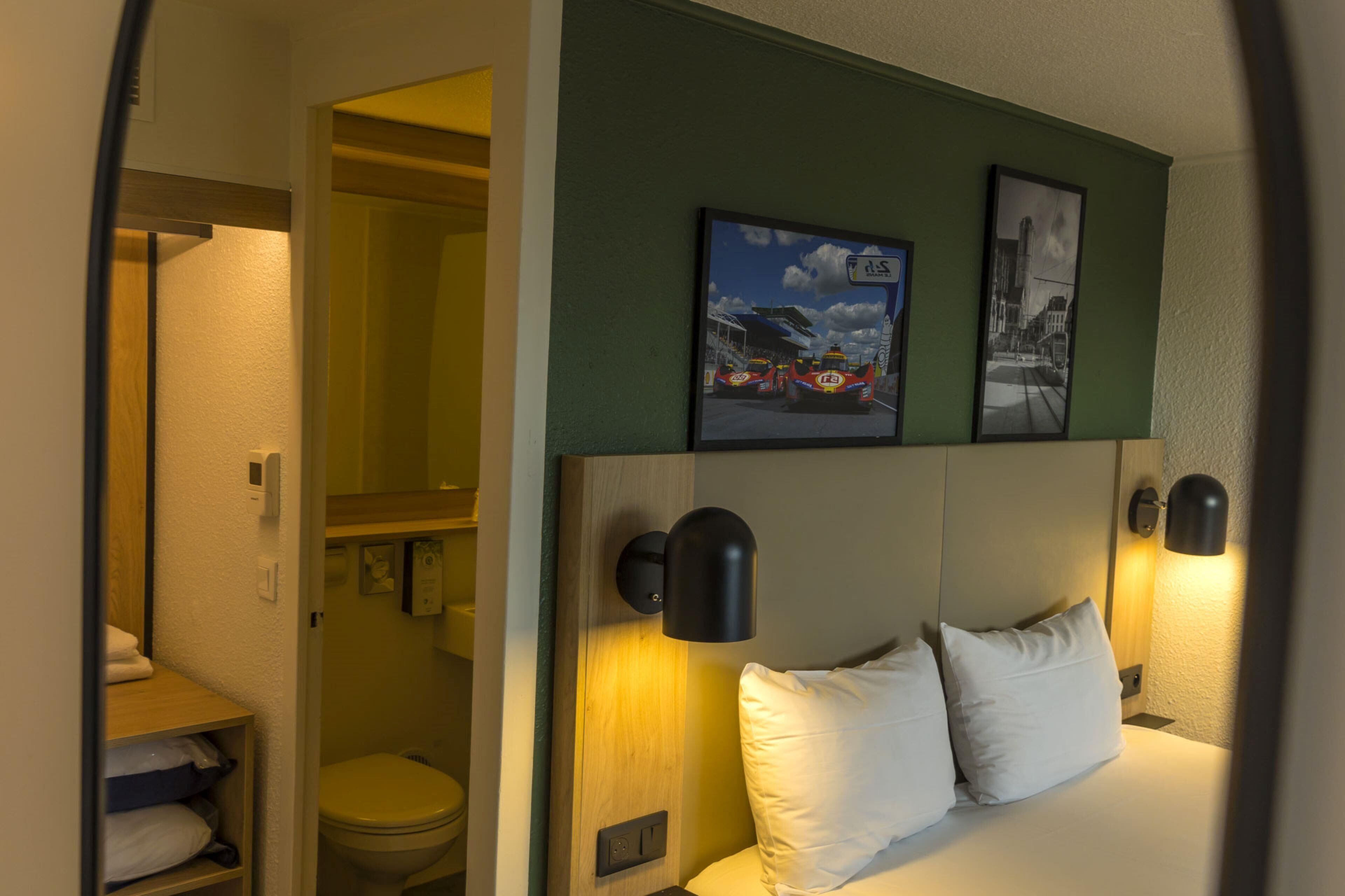 A hotel room with a bed, a headboard with wall-mounted lights, a toilet, and a shelf with towels.