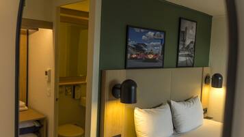 A hotel room with a bed, a headboard with wall-mounted lights, a toilet, and a shelf with towels.