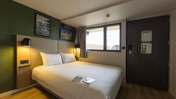 A hotel room with a large bed, a bedside table with a lamp, a window with curtains, and a door with a lock and a keycard slot.