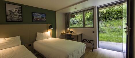 A hotel room with two beds, a desk with a chair, a window with a view of greenery, and a door leading to an outdoor area.
