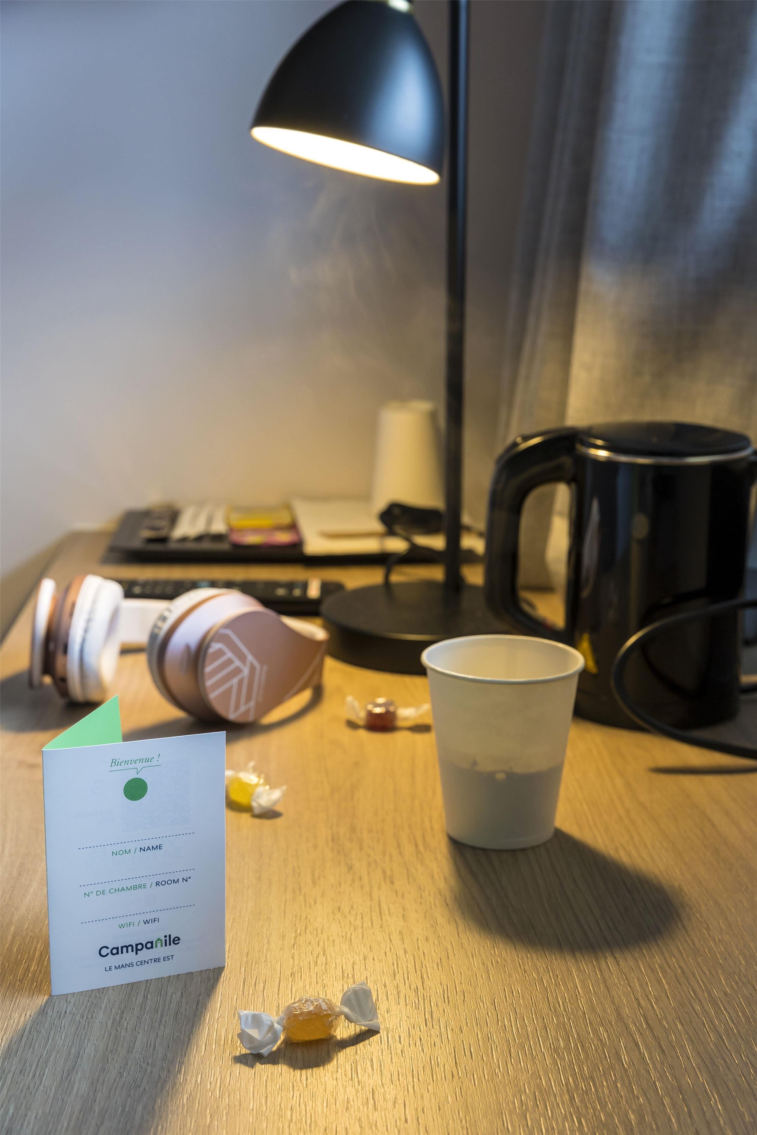 A hotel room with a wooden desk, a black lamp, a white cup, a pair of headphones, a coffee maker, and a welcome card.