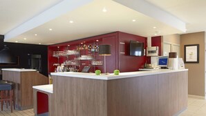 A modern bar area with a wooden counter, shelves with bottles and glasses, and a digital screen. - Campanile NATURE - Le Mans Est (Le Mans)
