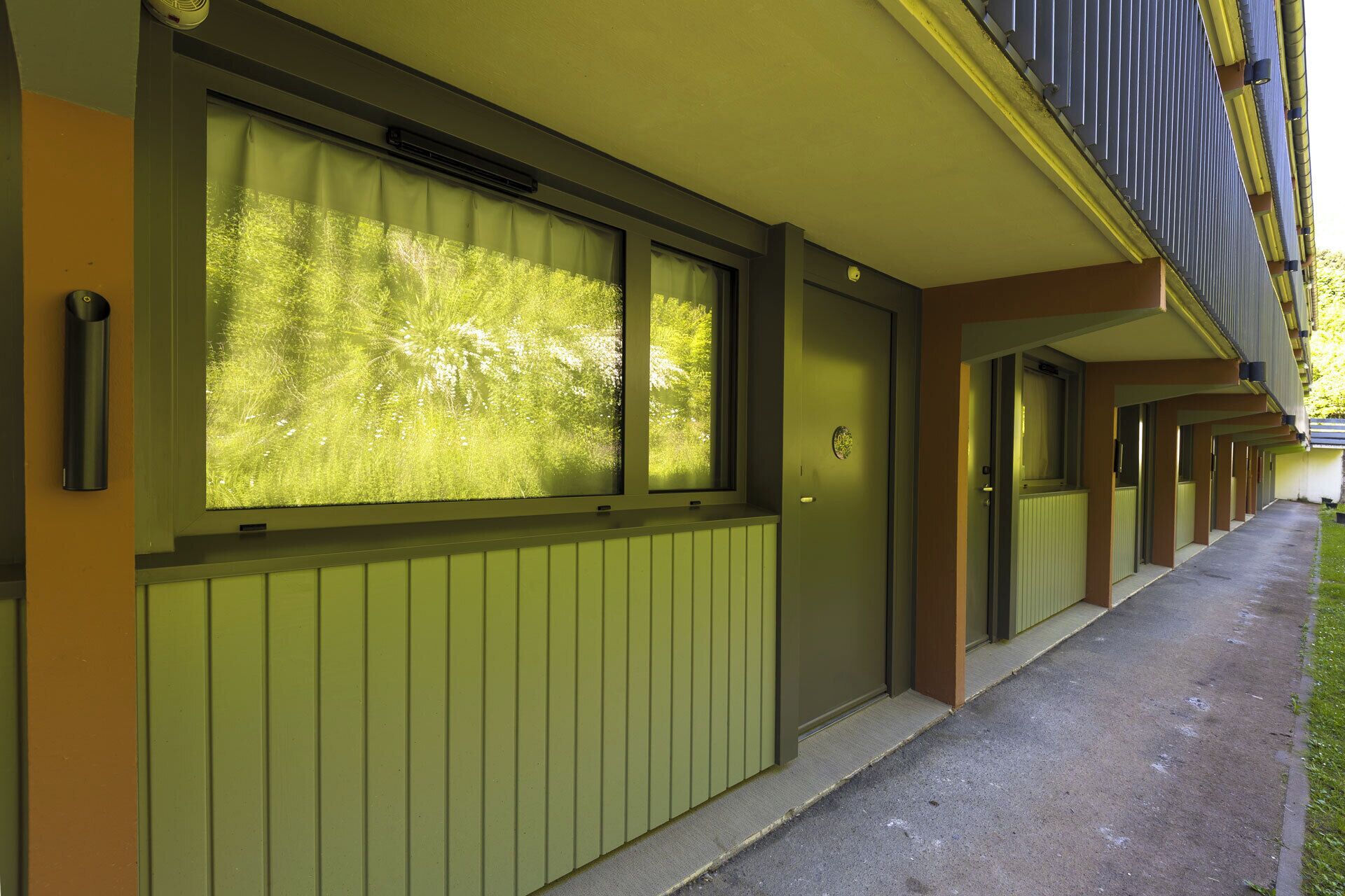 A row of hotel rooms with large windows, a green exterior, and a covered walkway.