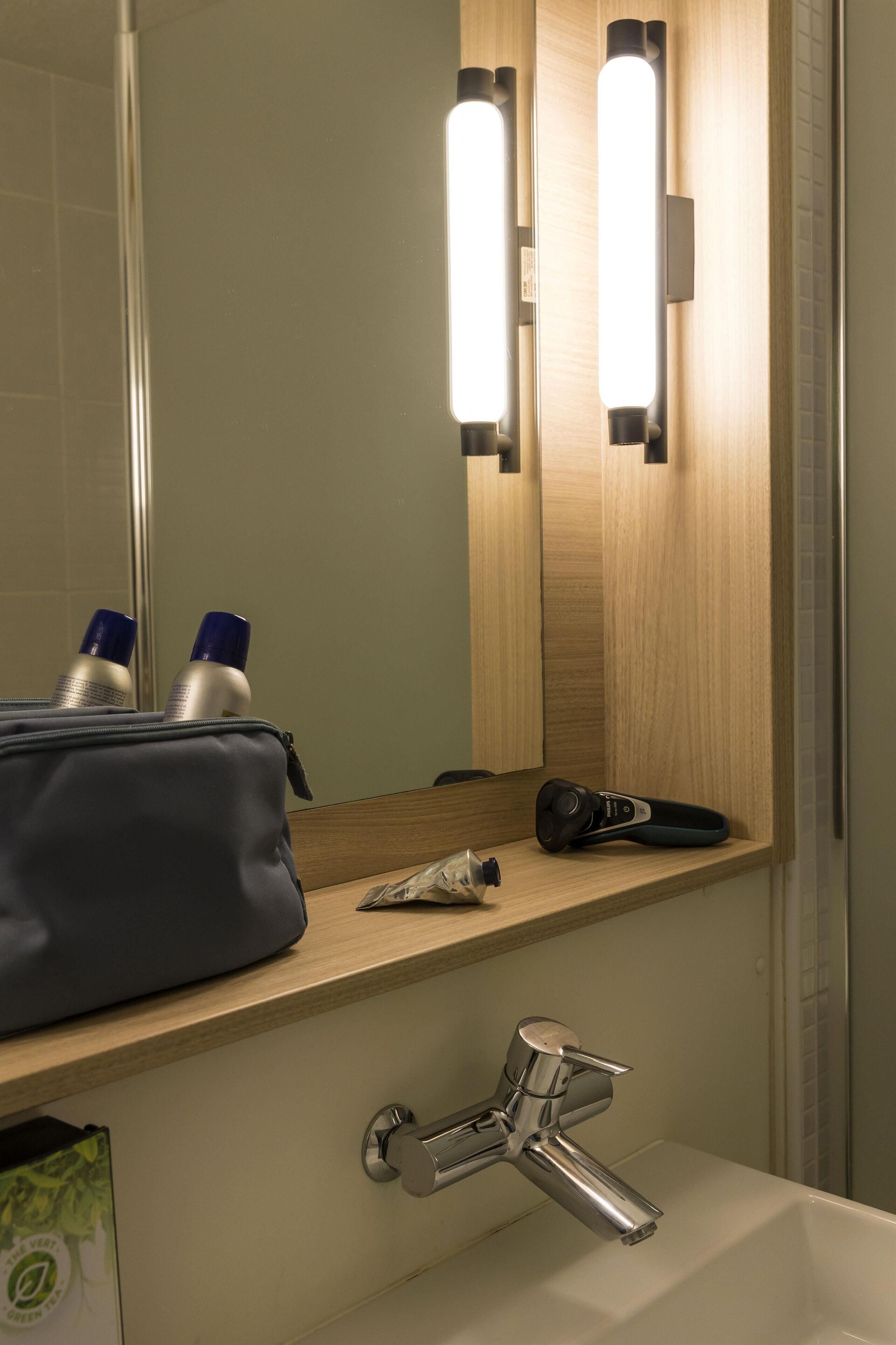 Une salle de bain avec un miroir, deux appliques murales, un lavabo avec un robinet et une étagère contenant des articles de toilette.