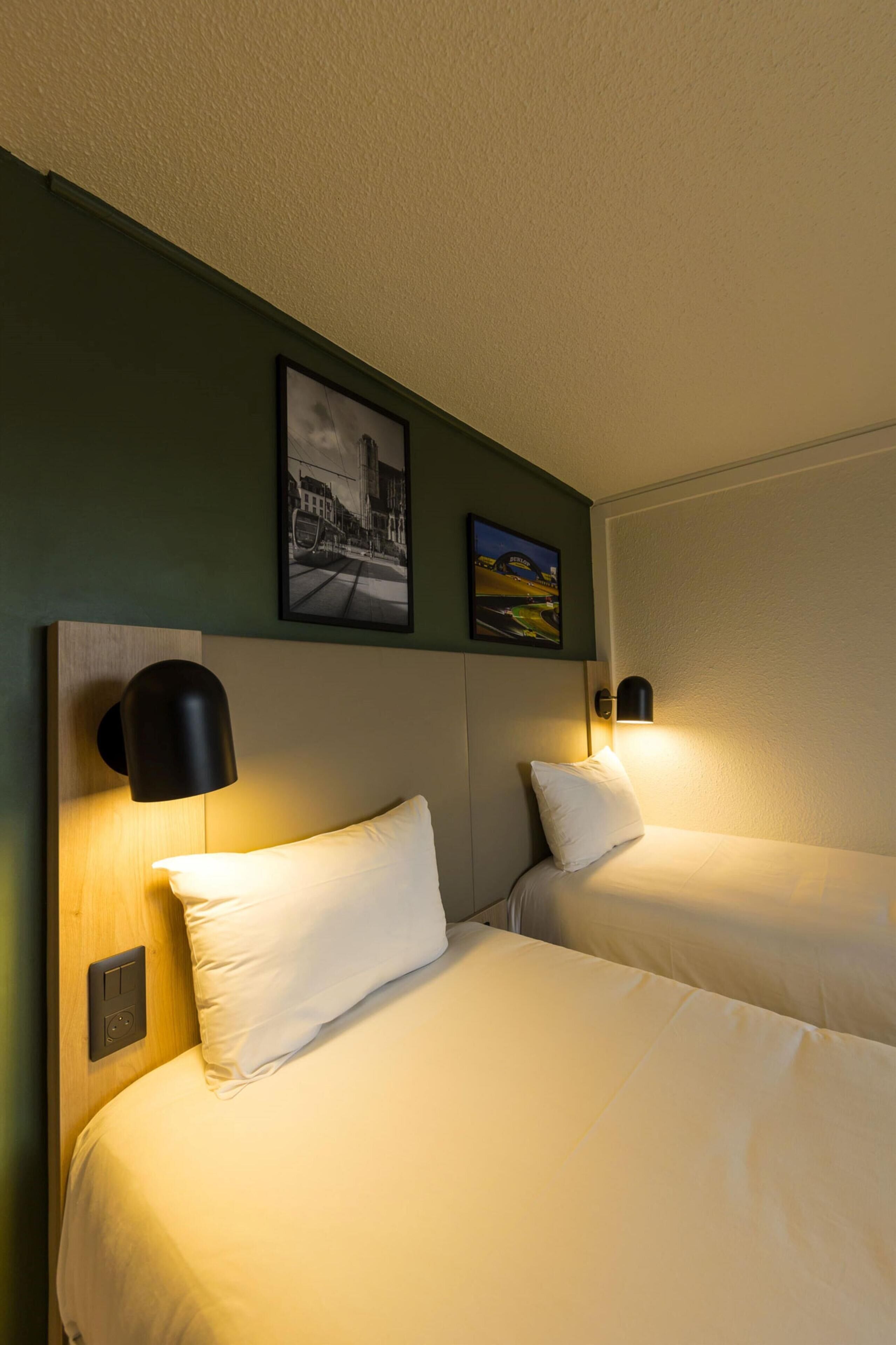 Habitación de hotel con dos camas, cada una con un almohadón blanco y una lámpara negra de pared. Encima de las camas hay un cuadro enmarcado.