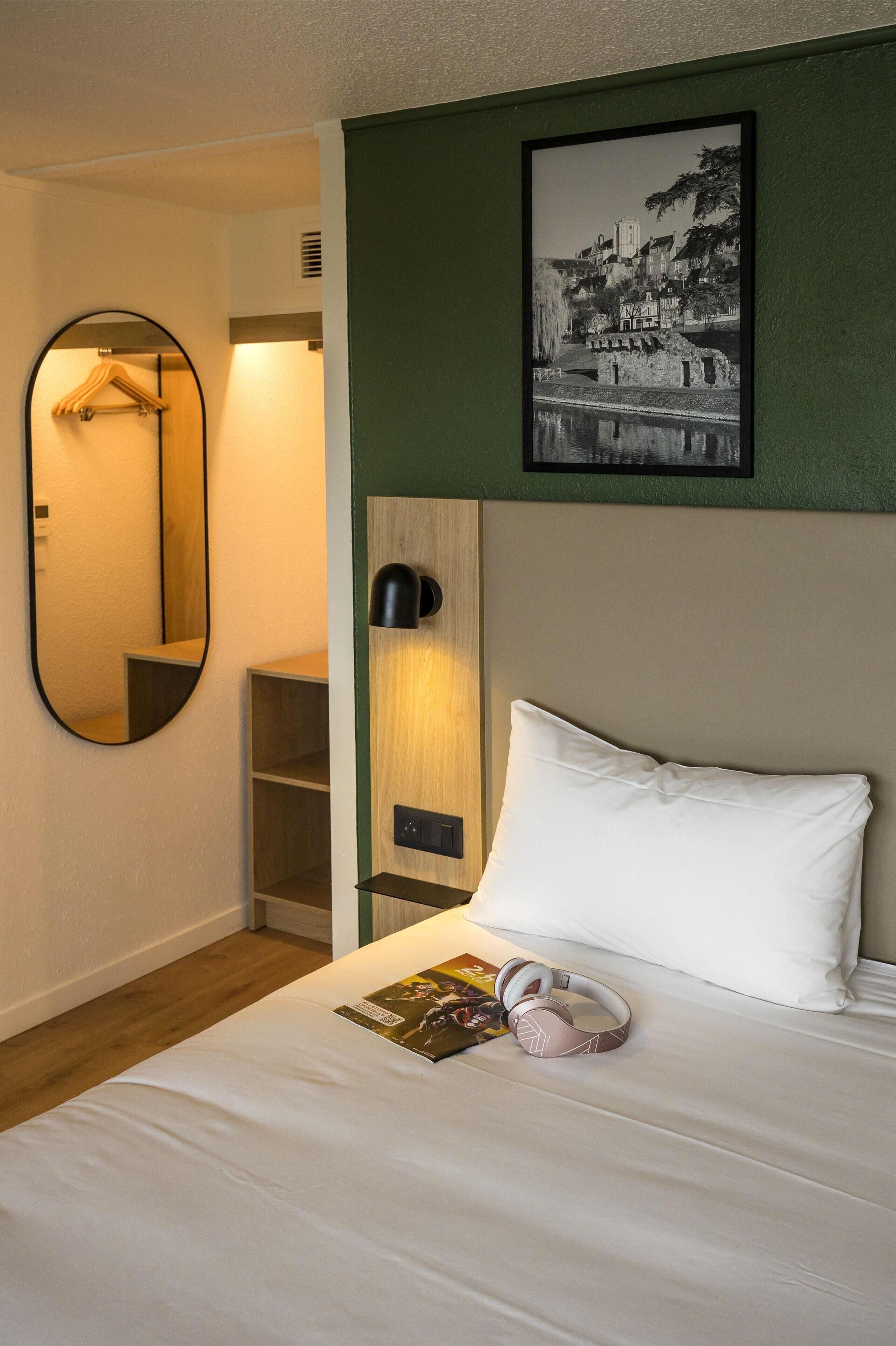 A hotel room with a bed, a mirror, a shelf, a framed picture, and a pillow.
