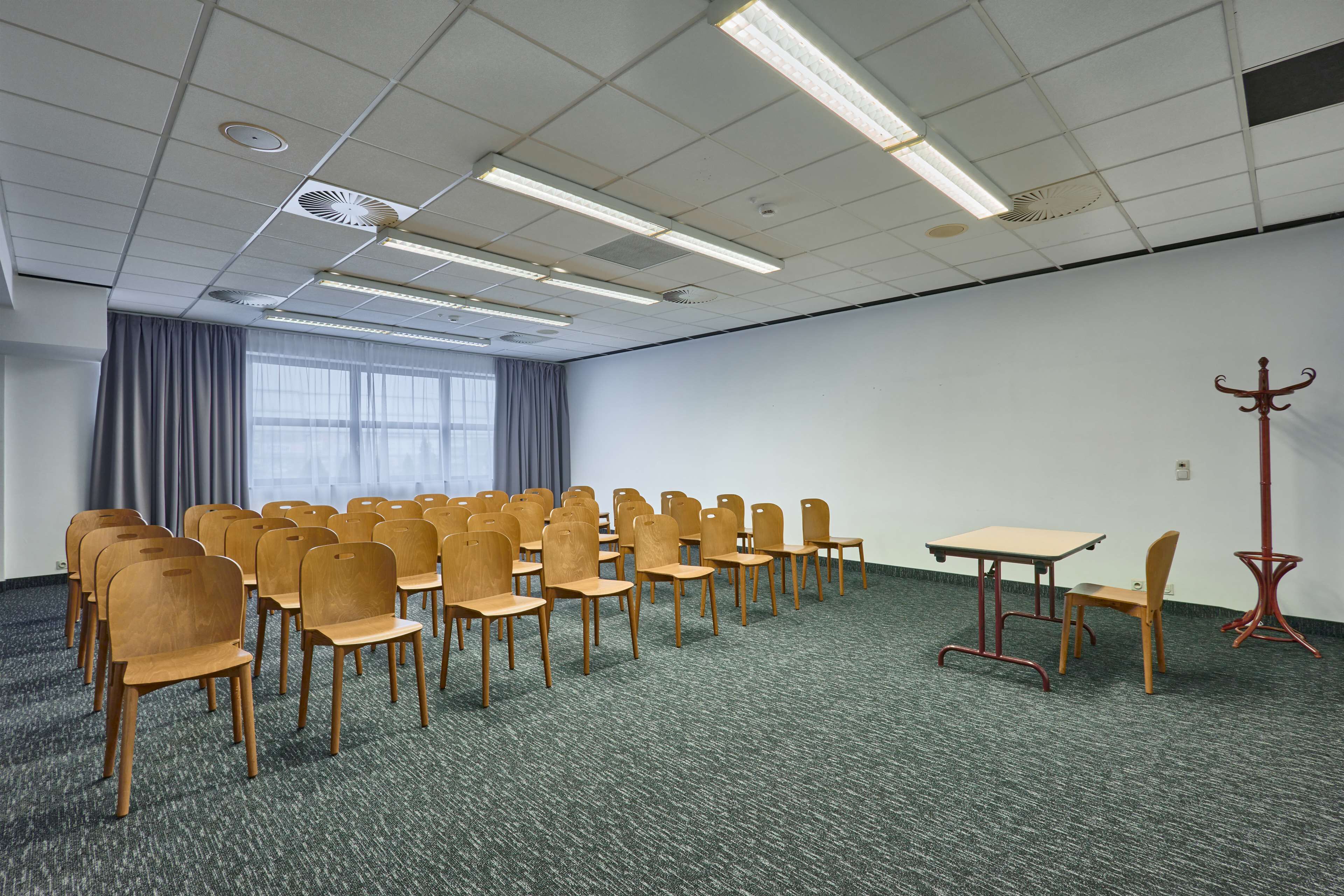 Een vergaderzaal met rijen houten stoelen, een tafel en een rode vloerlamp.
