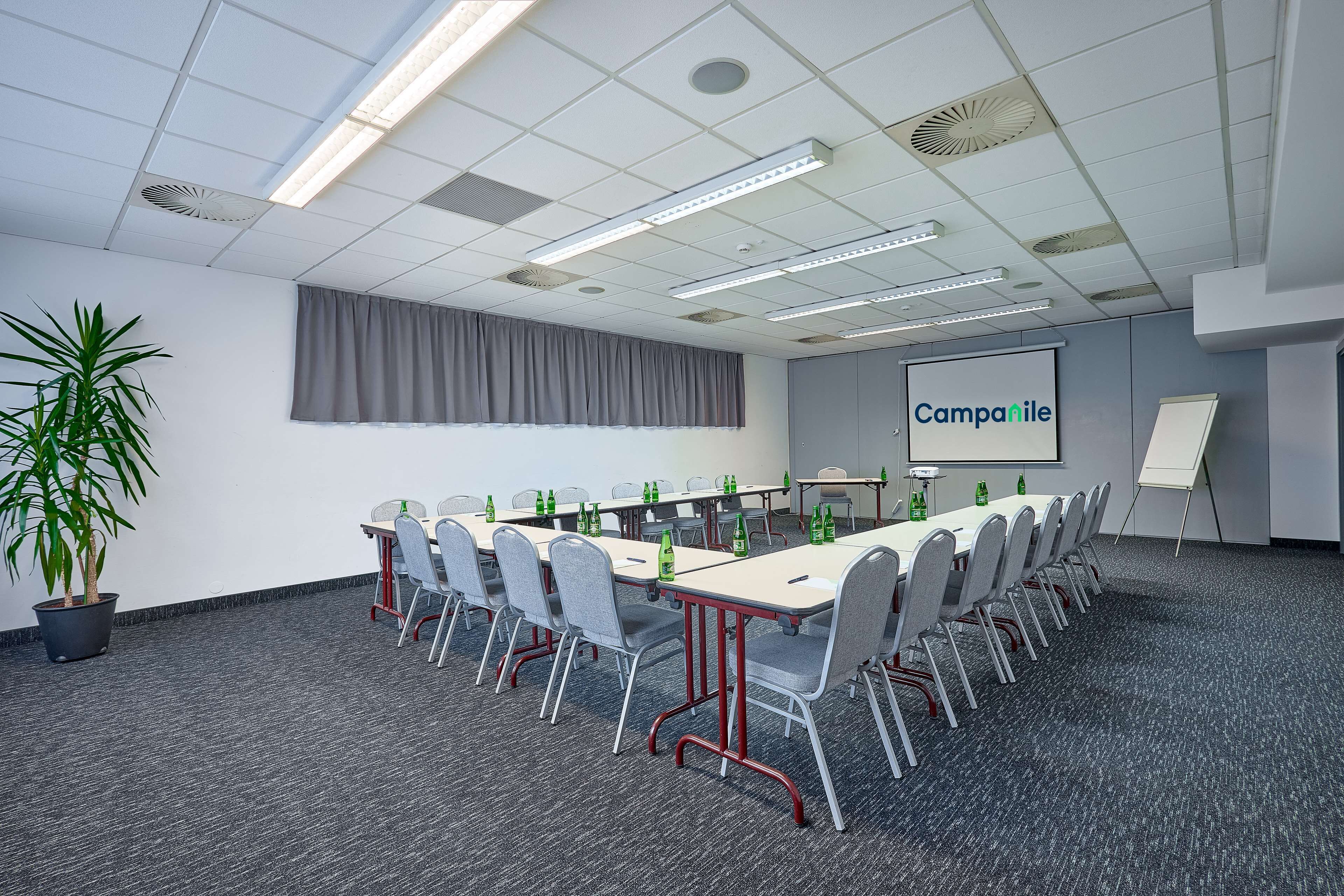 A conference room with a long table, chairs, a projector screen, and a potted plant.