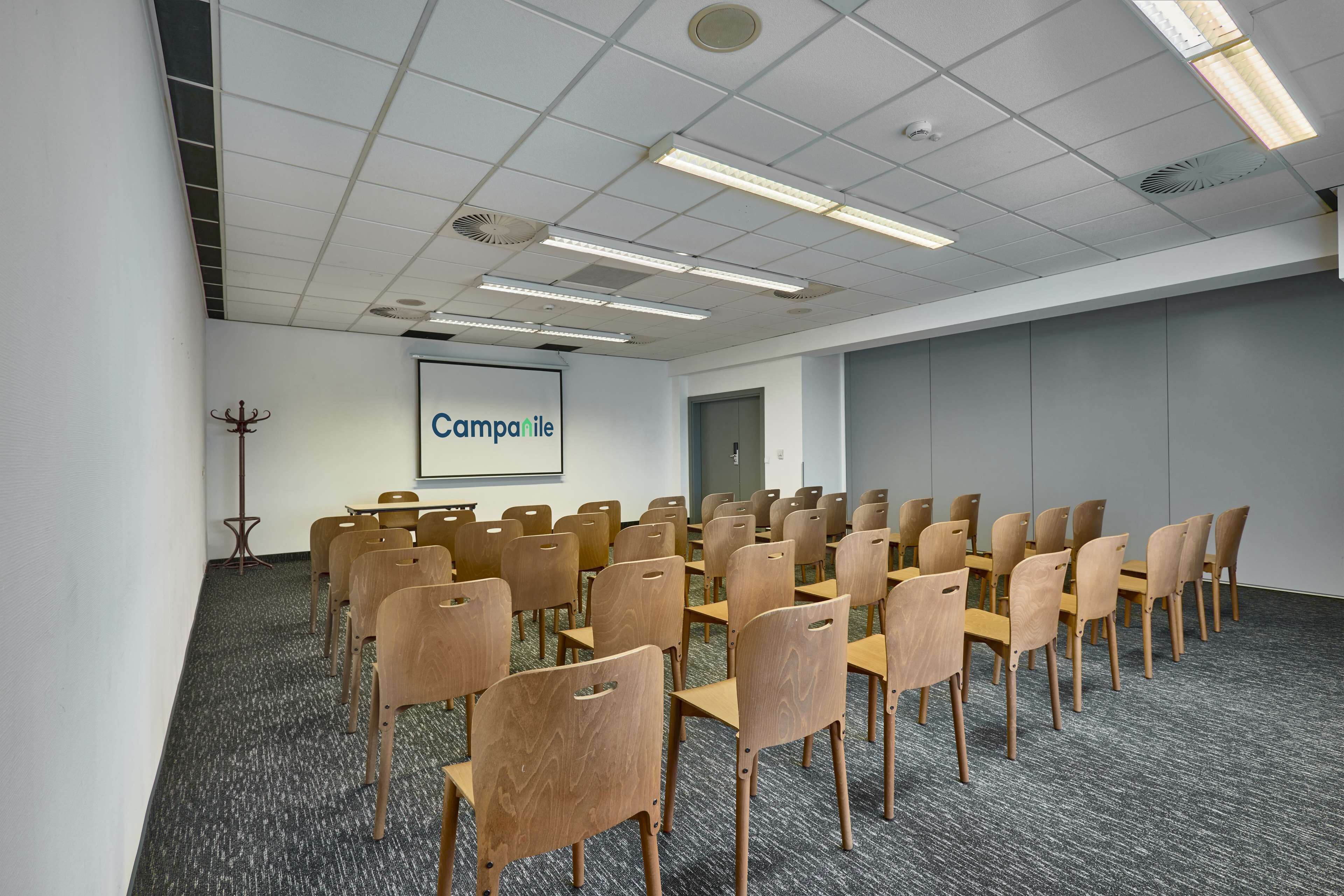 A conference room with rows of wooden chairs and tables, a projector screen with "Campanile" text, and a grey carpeted floor.