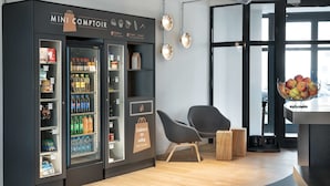 A modern interior with a vending machine labeled 'Mini Comptoir' stocked with beverages and snacks, a wooden counter, and a seating area with chairs. - Campanile Katowice (Katowice)
