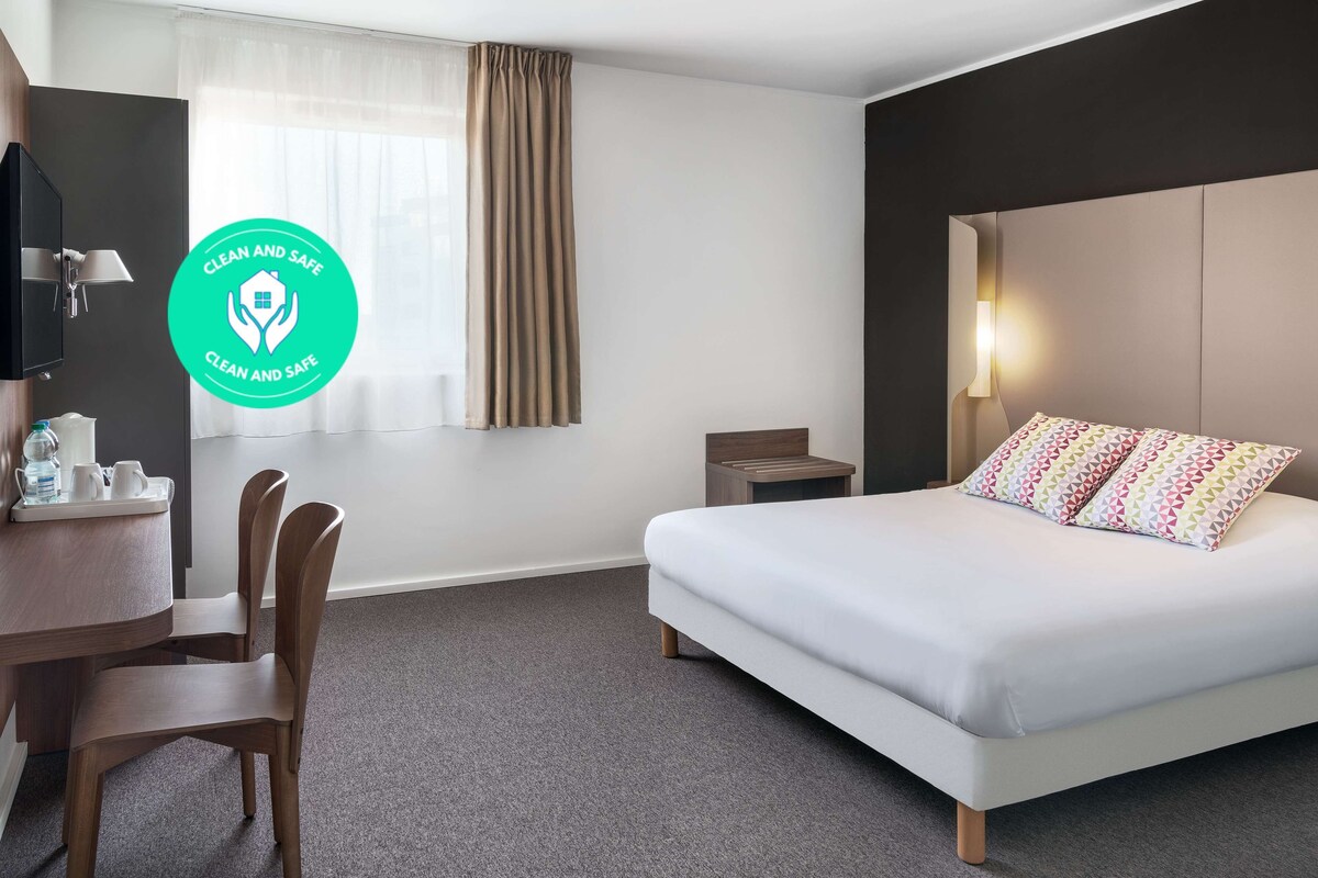 A hotel room with a bed, a desk, chairs, a nightstand, and a window with curtains.