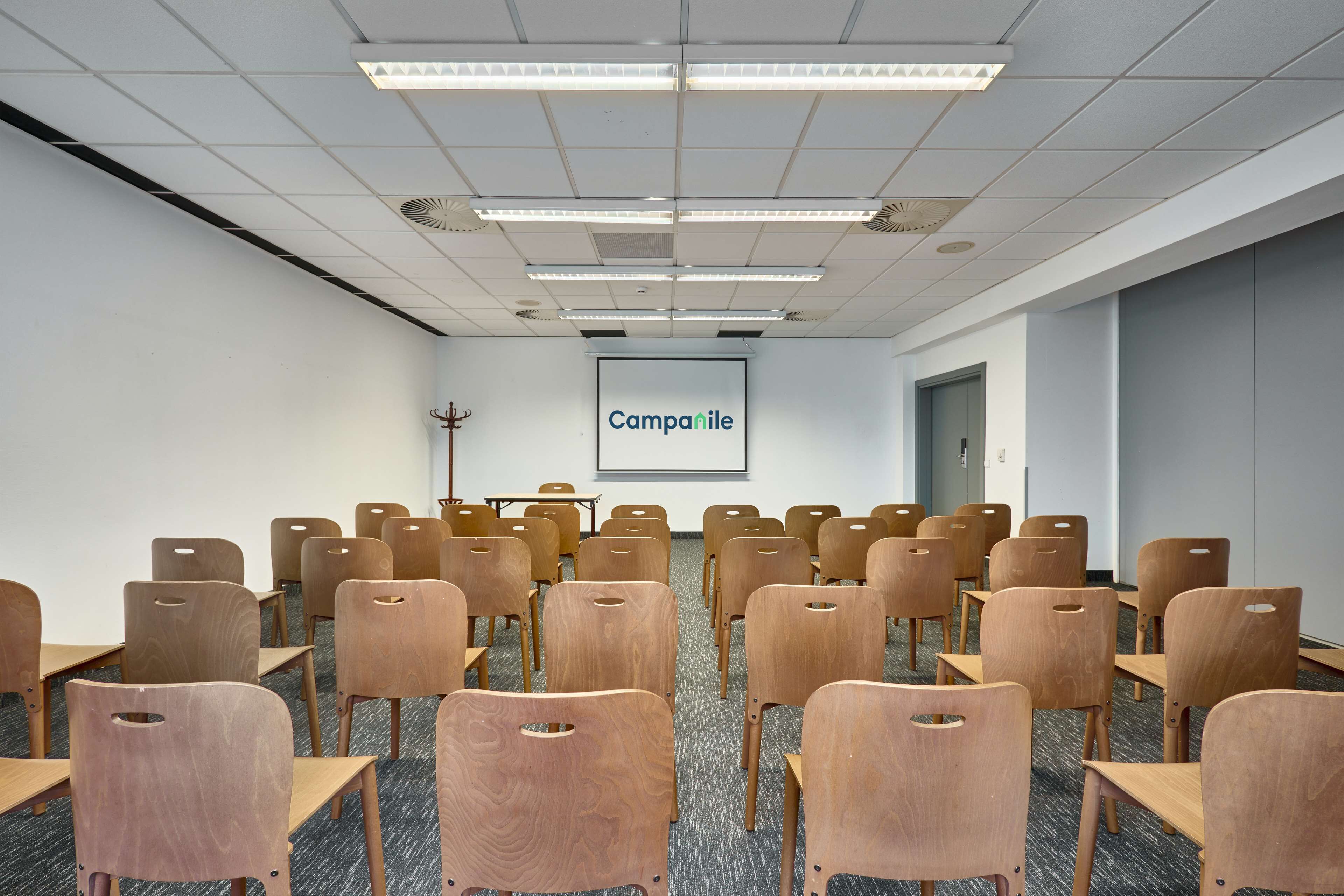 A conference room with rows of wooden chairs and tables, a projector screen with the word "Campanile," and a door in the background.