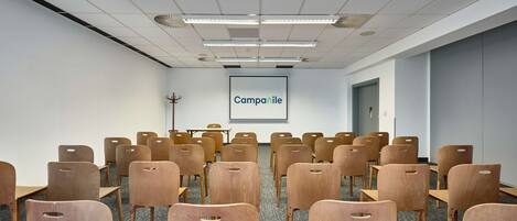 A conference room with rows of wooden chairs and tables, a projector screen with the word "Campanile," and a door in the background.