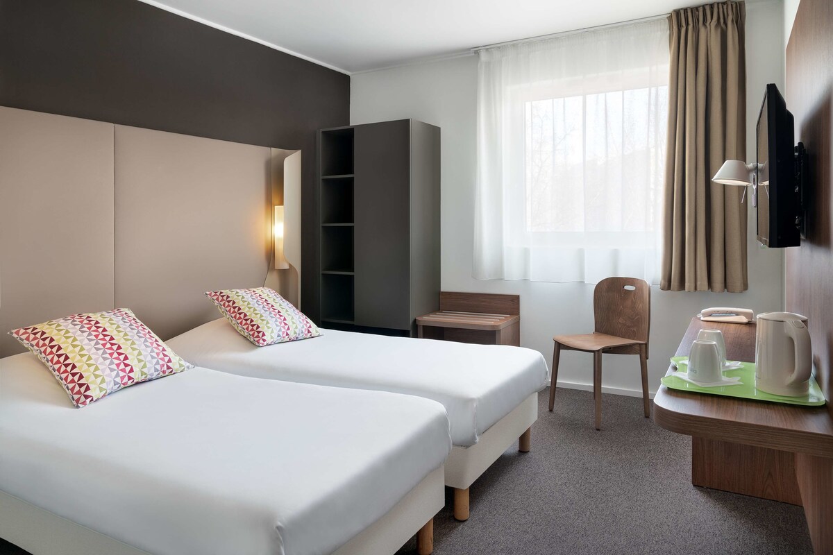 A hotel room with two beds, a desk with a coffee maker, a chair, a TV mounted on the wall, and a window with curtains.