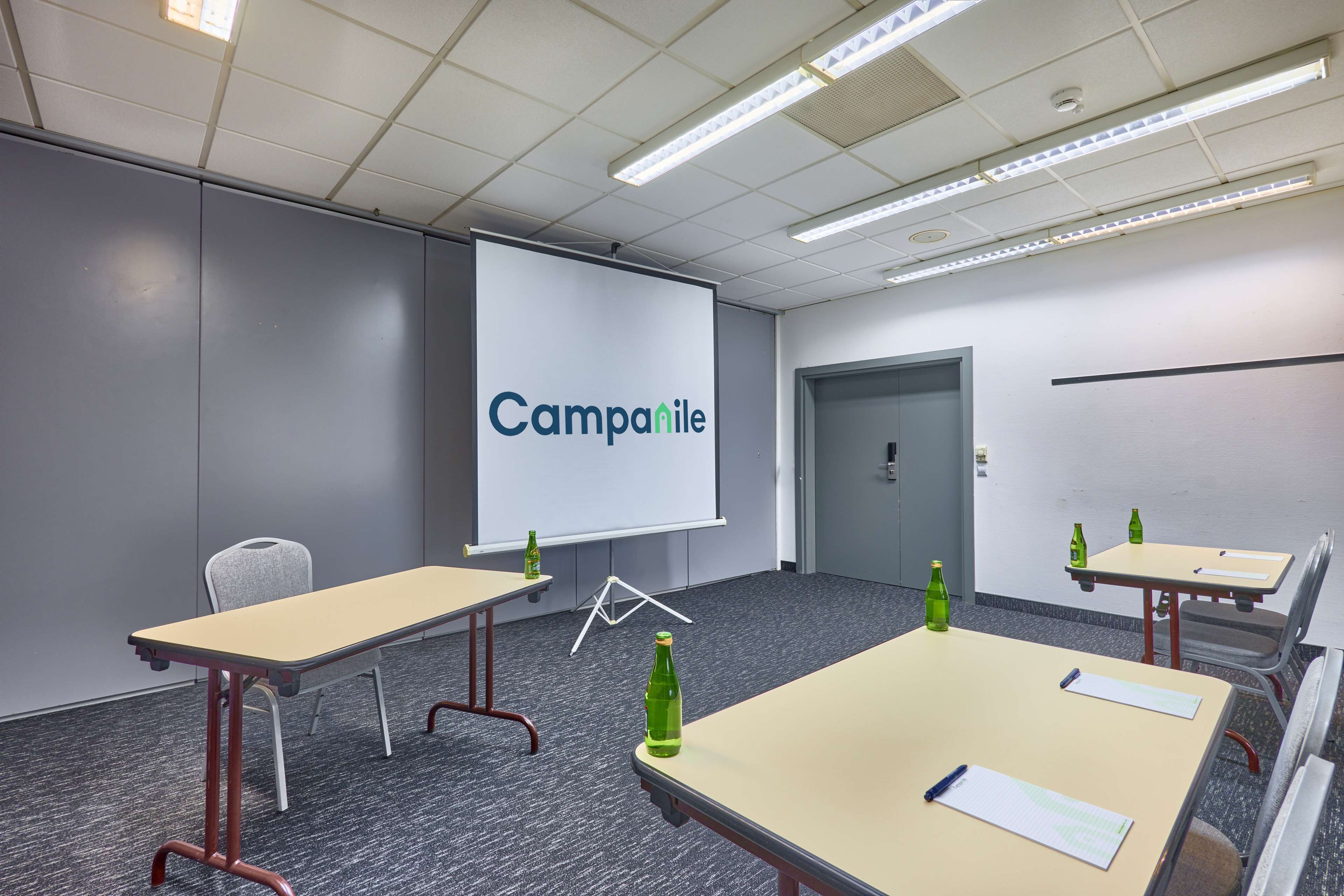 Een vergaderruimte met een projectiescherm waarop ‘Campanile’ staat, tafels met stoelen en flessen op de tafels.