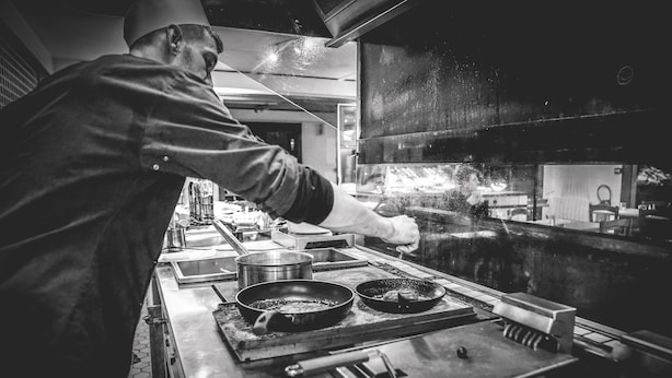Um chef cozinha em uma cozinha profissional com superfícies de aço inoxidável, panelas, frigideiras e um forno grande.
