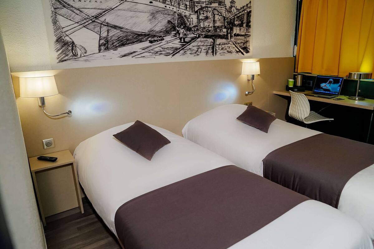Een hotelkamer met twee bedden, elk met een kussen en een bedlampje. Er is een bureau met een computer en een lamp, en een kunstwerk aan de muur boven de bedden.