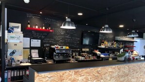 Uma área de bar com balcão de madeira, banquetas, máquina de café e uma televisão na parede.