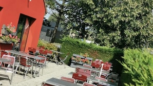 Uma área externa com mesas e cadeiras, um edifício vermelho e um arbusto ao fundo.