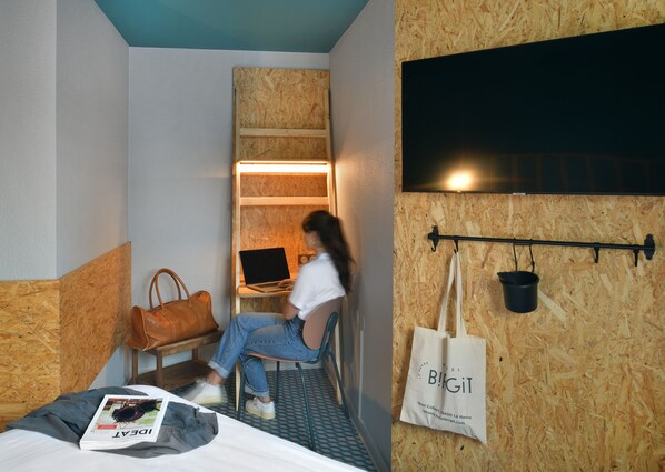 Habitación de hotel pequeña con cama, escritorio con computadora portátil, silla y televisor montado en la pared.