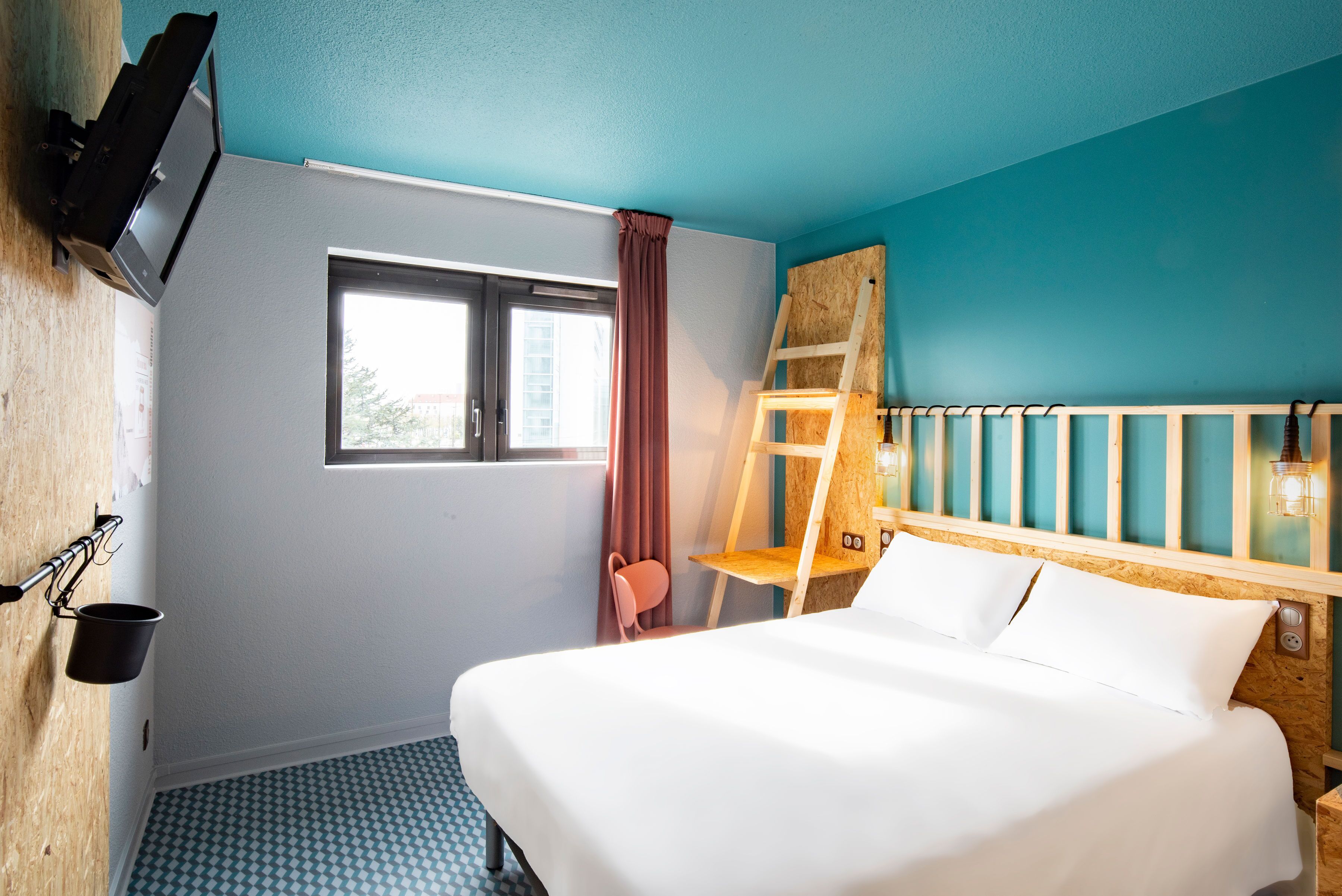 A hotel room with a bed, a window with curtains, a wooden ladder, a small table, and a TV mounted on the wall.