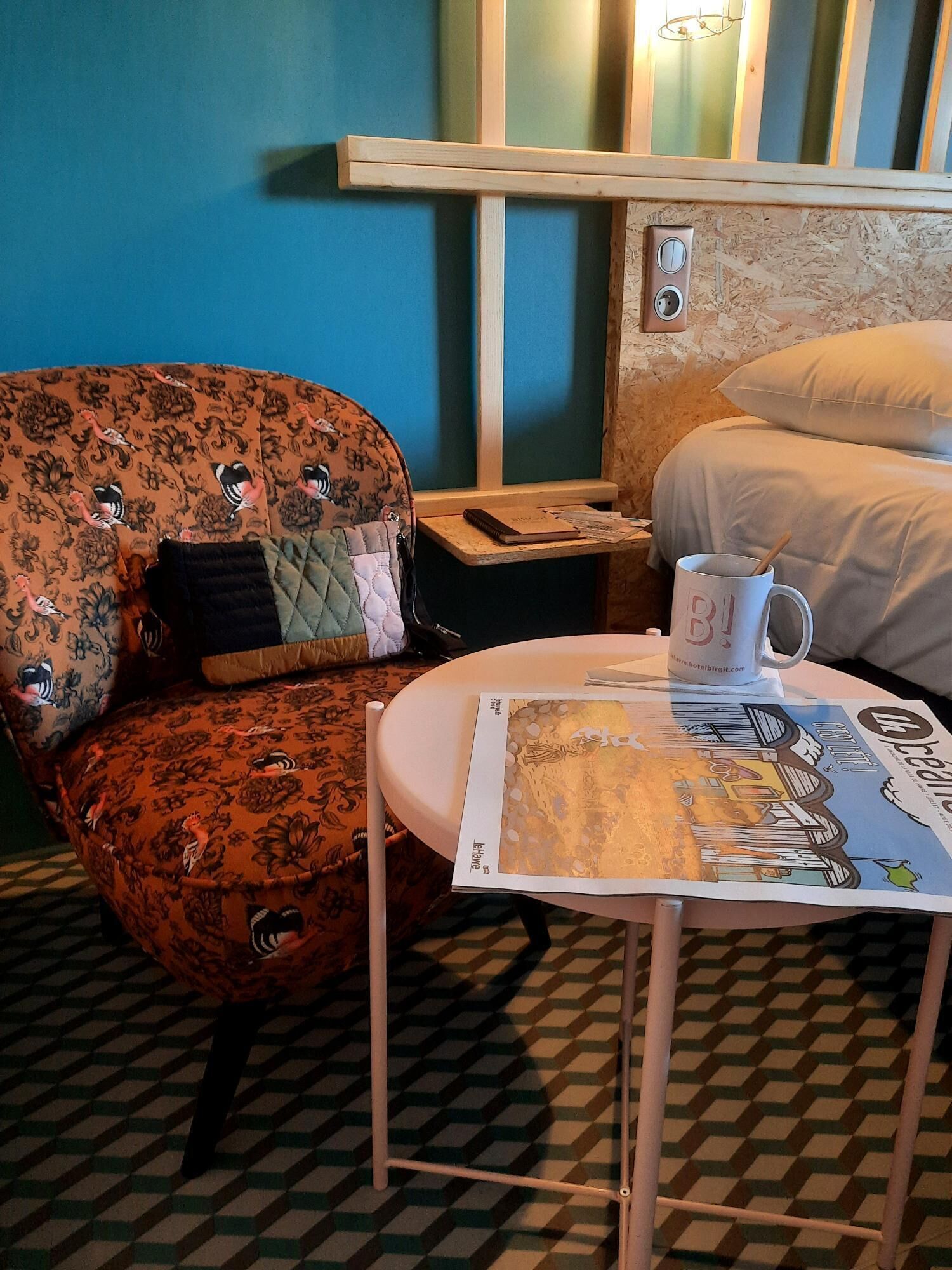 A hotel room with a floral-patterned armchair, a small table with a magazine, a white bed, and a blue wall.