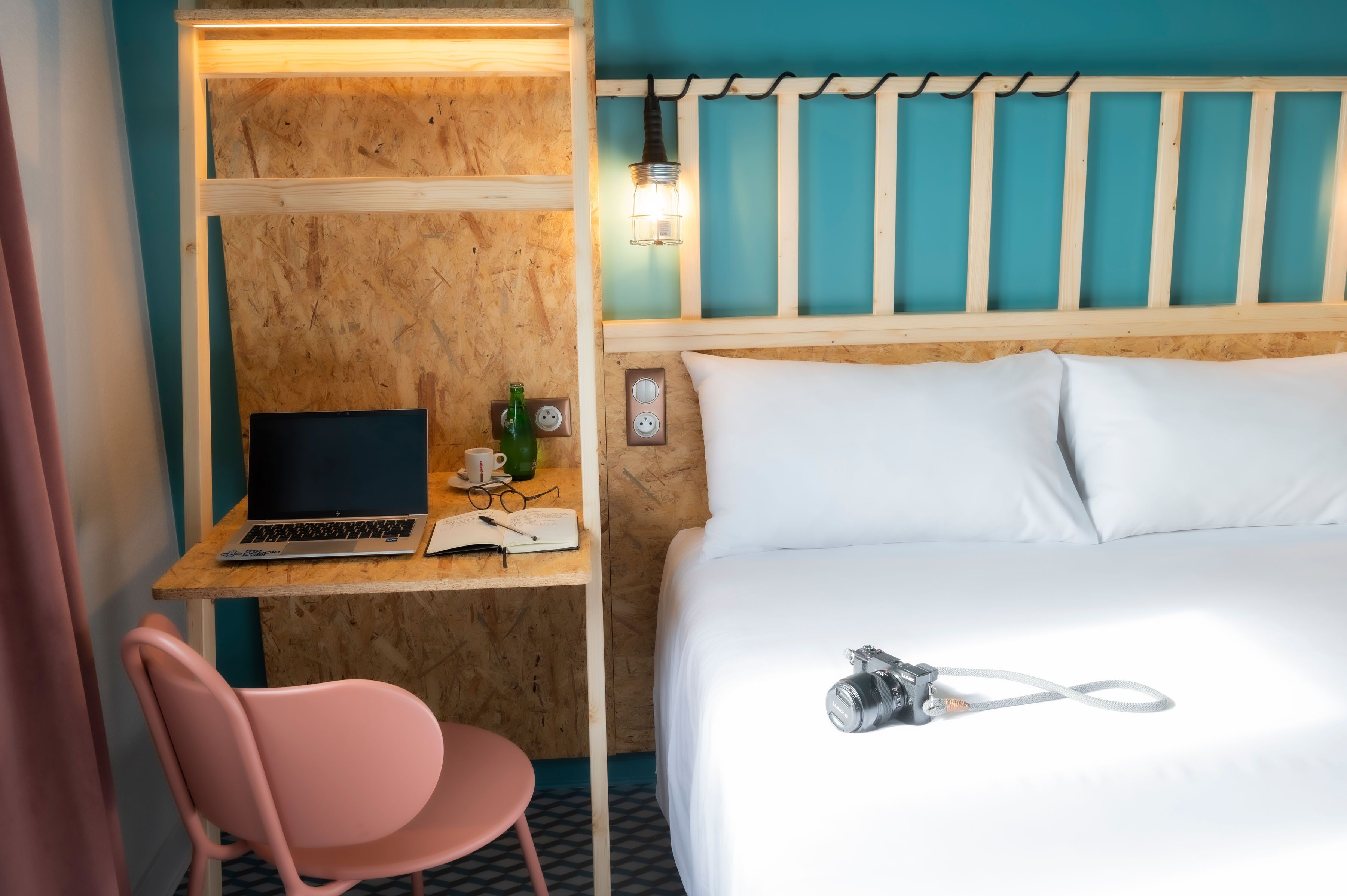 Hotellihuone, jossa on puinen työpöytä, kannettava tietokone, vaaleanpunainen tuoli, valkoisilla lakanoilla varustettu sänky ja kamera sängyllä.