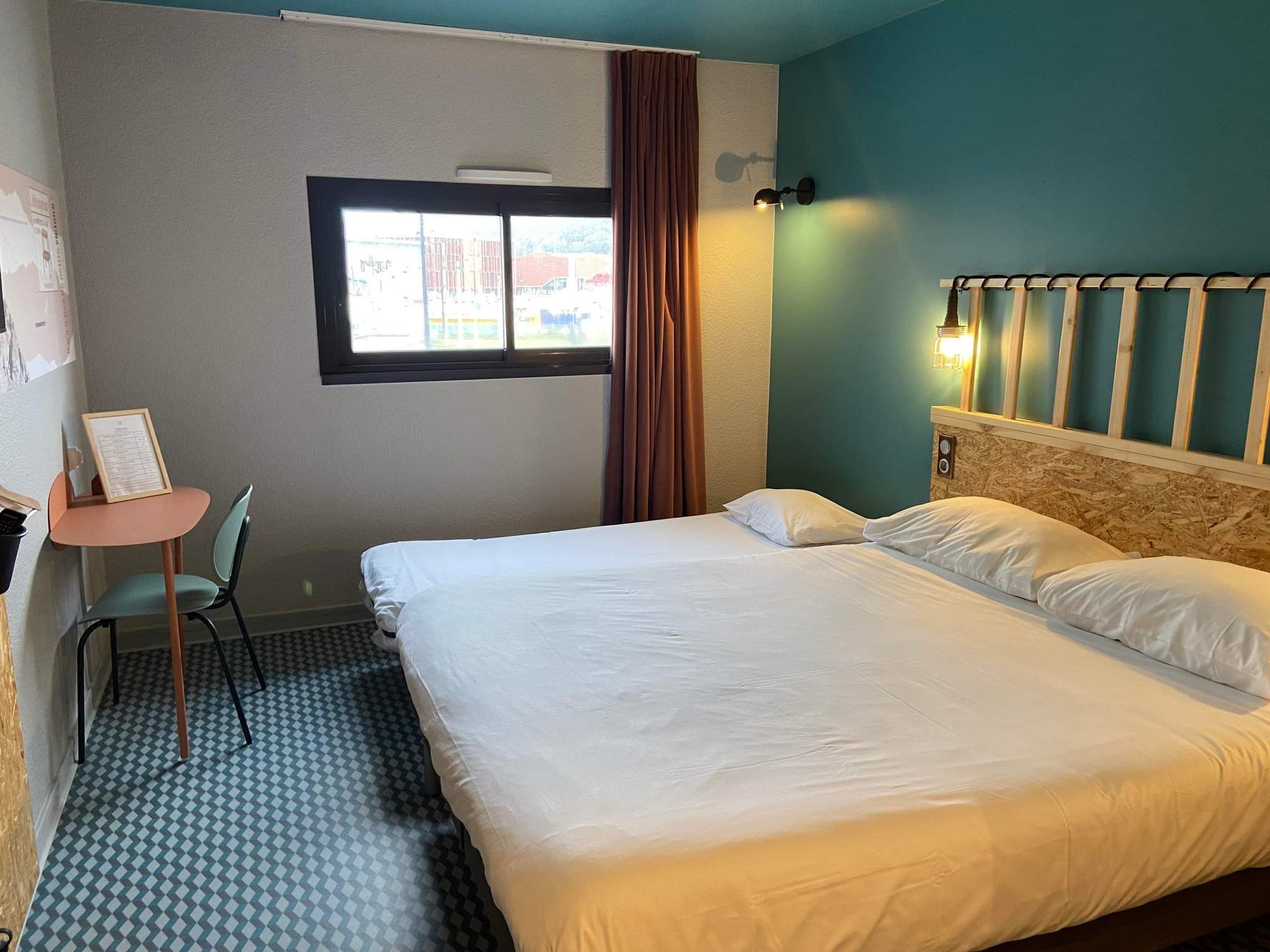 Habitación de hotel con cama, escritorio, silla, ventana y lámpara fijada a la pared.