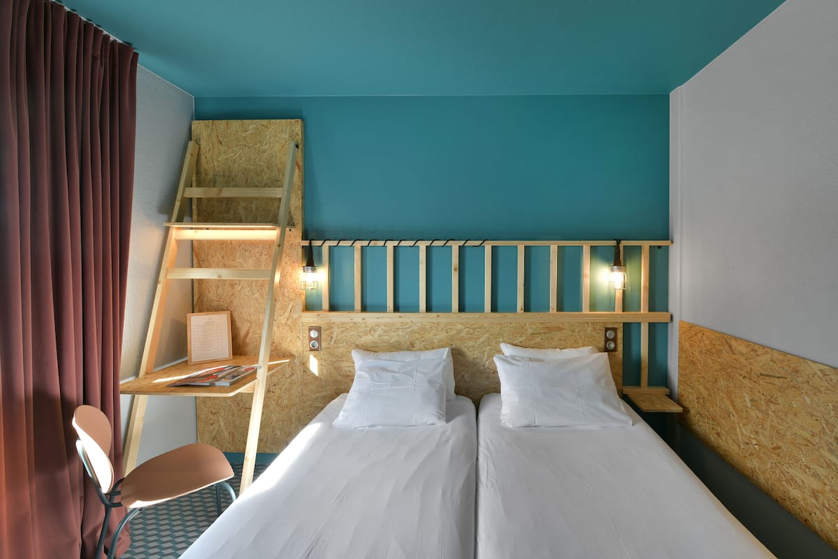 A hotel room with two beds, a wooden ladder, a desk with a chair, and a window with curtains.