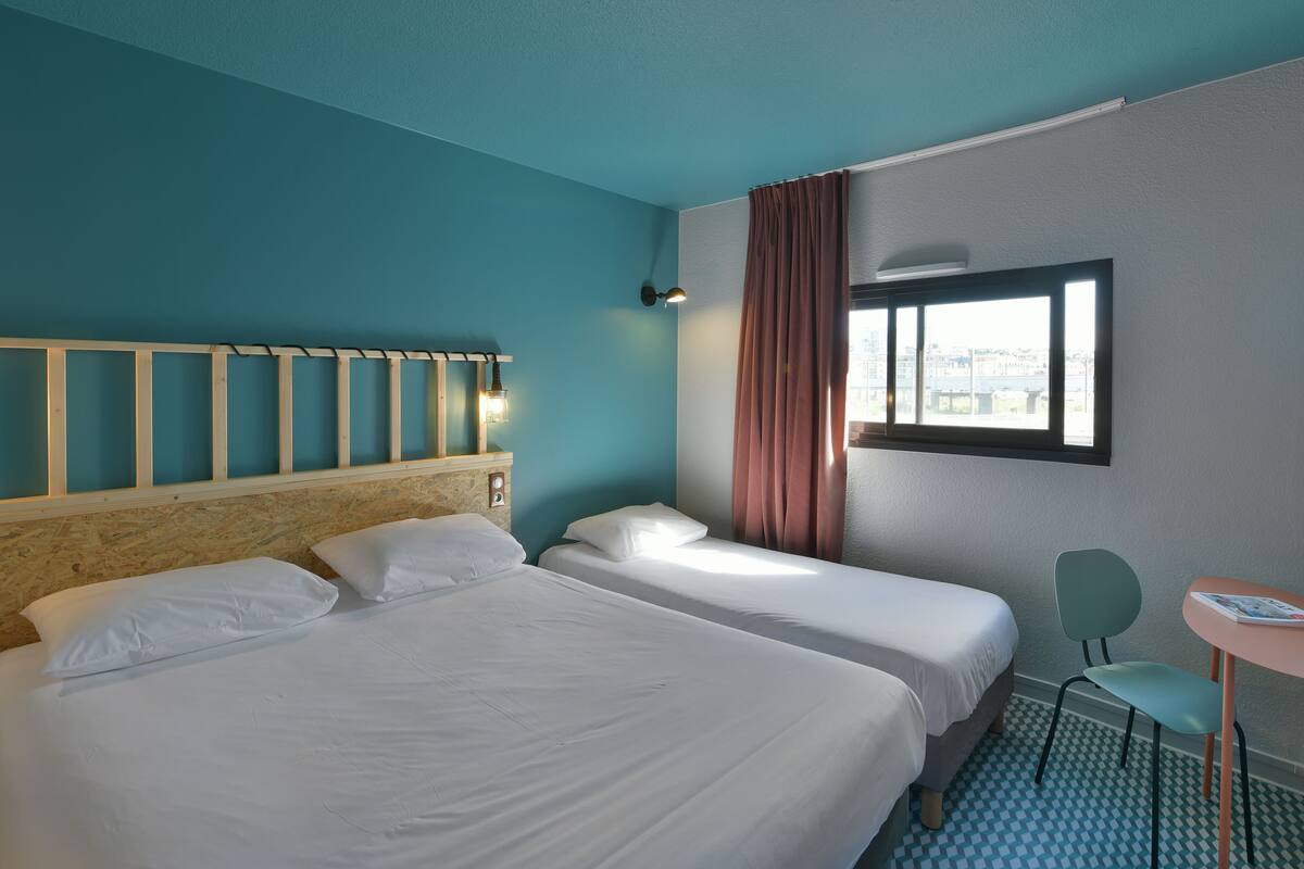A hotel room with two beds, a window with curtains, a small table with a chair, and a view of the cityscape.