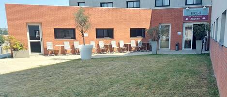 A building with a red brick facade, a sign for 'BIRGIIT' with a contact number, and a seating area with chairs and a planter.