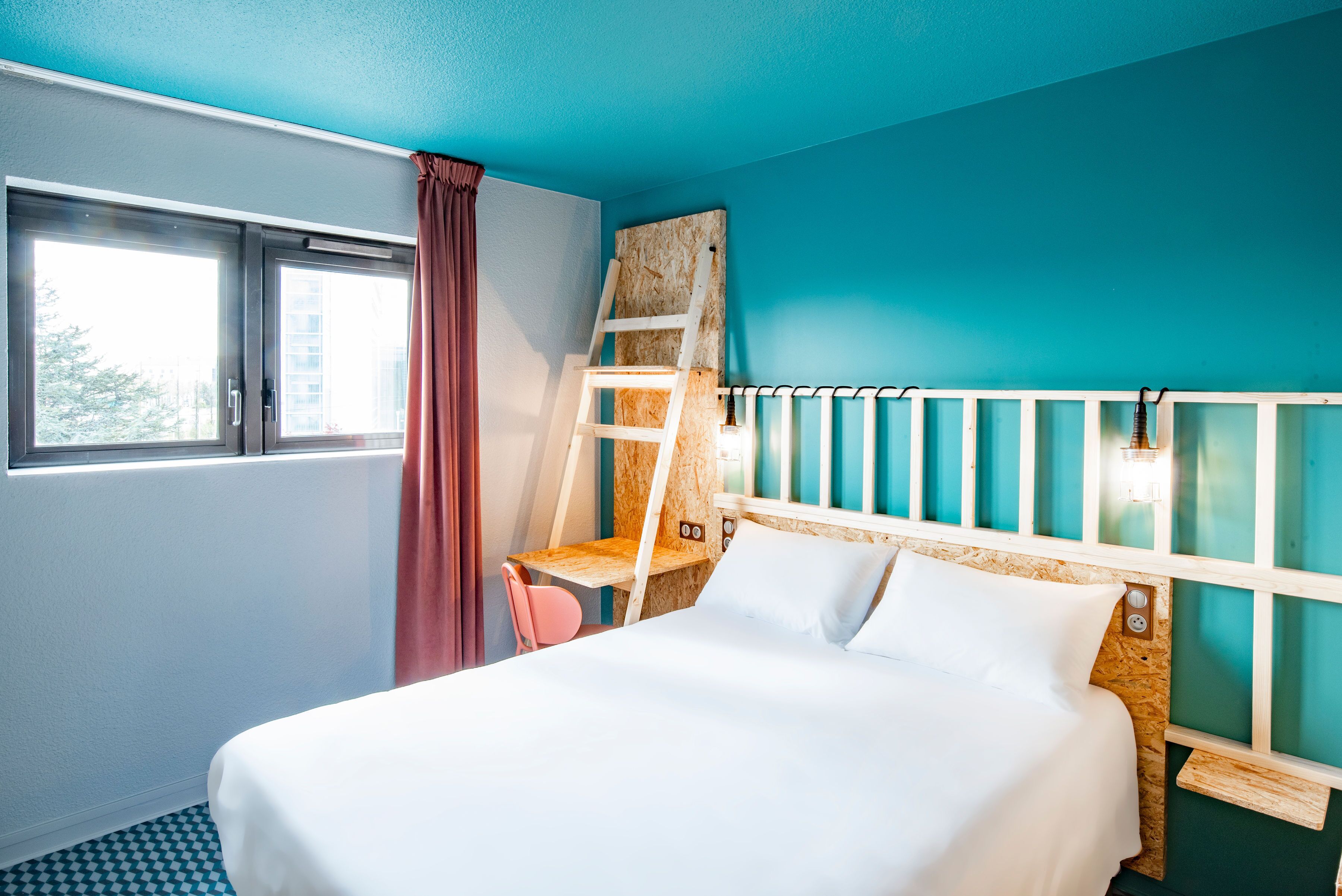 A hotel room with a bed, a desk, a chair, a window with curtains, and a view of the outside.
